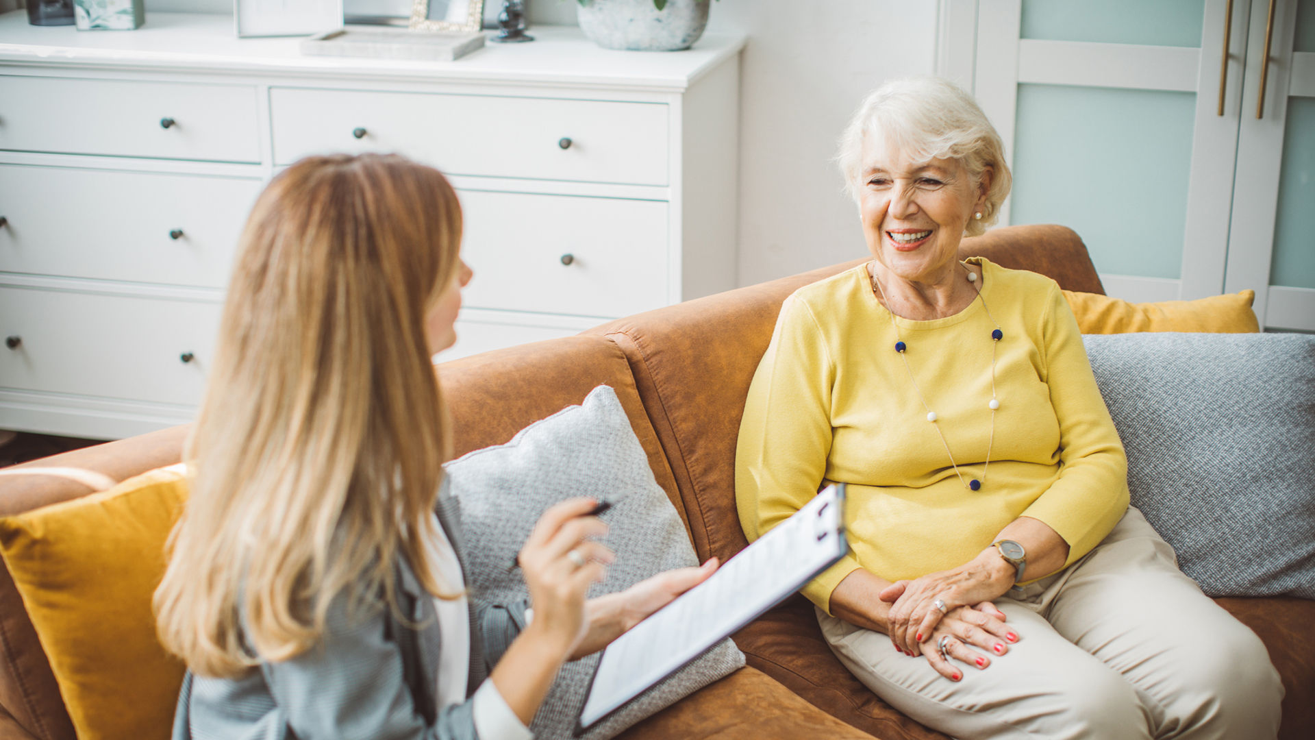 Sales counselor meeting with a senior prospective resident on orange couch in apartment.