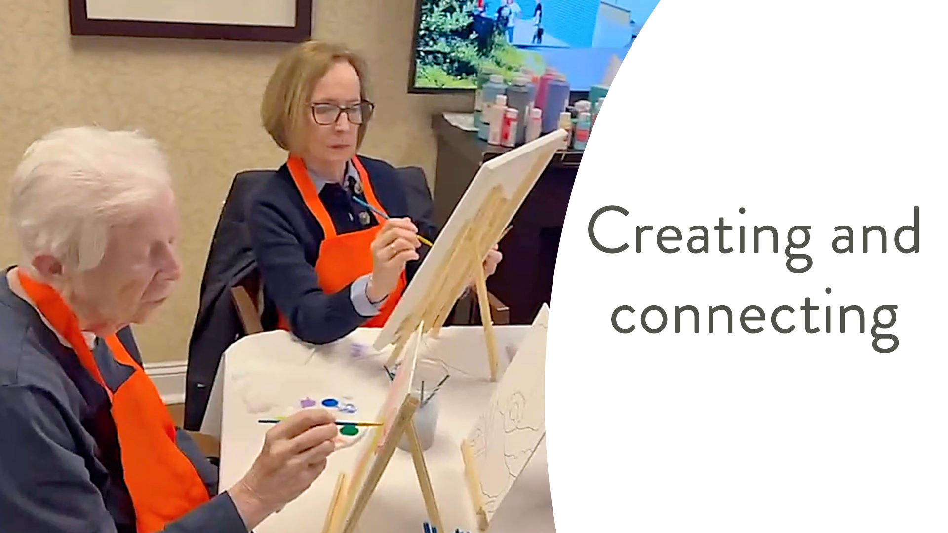 Two people wearing orange aprons are painting on canvases at a table. The text on the image reads, "Creating and connecting."