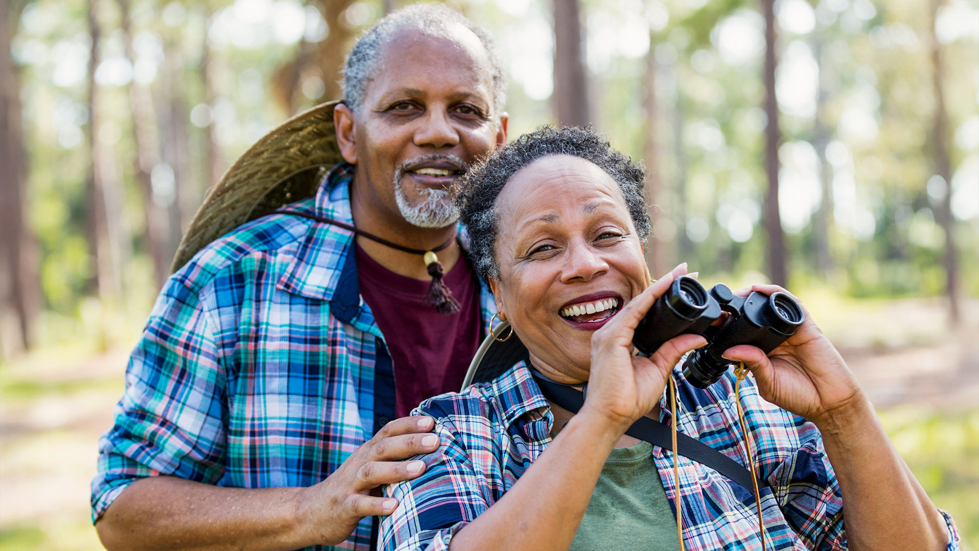 Smiling couple in plaid shirts, with the woman holding binoculars, enjoying a day in a sunlit forest.