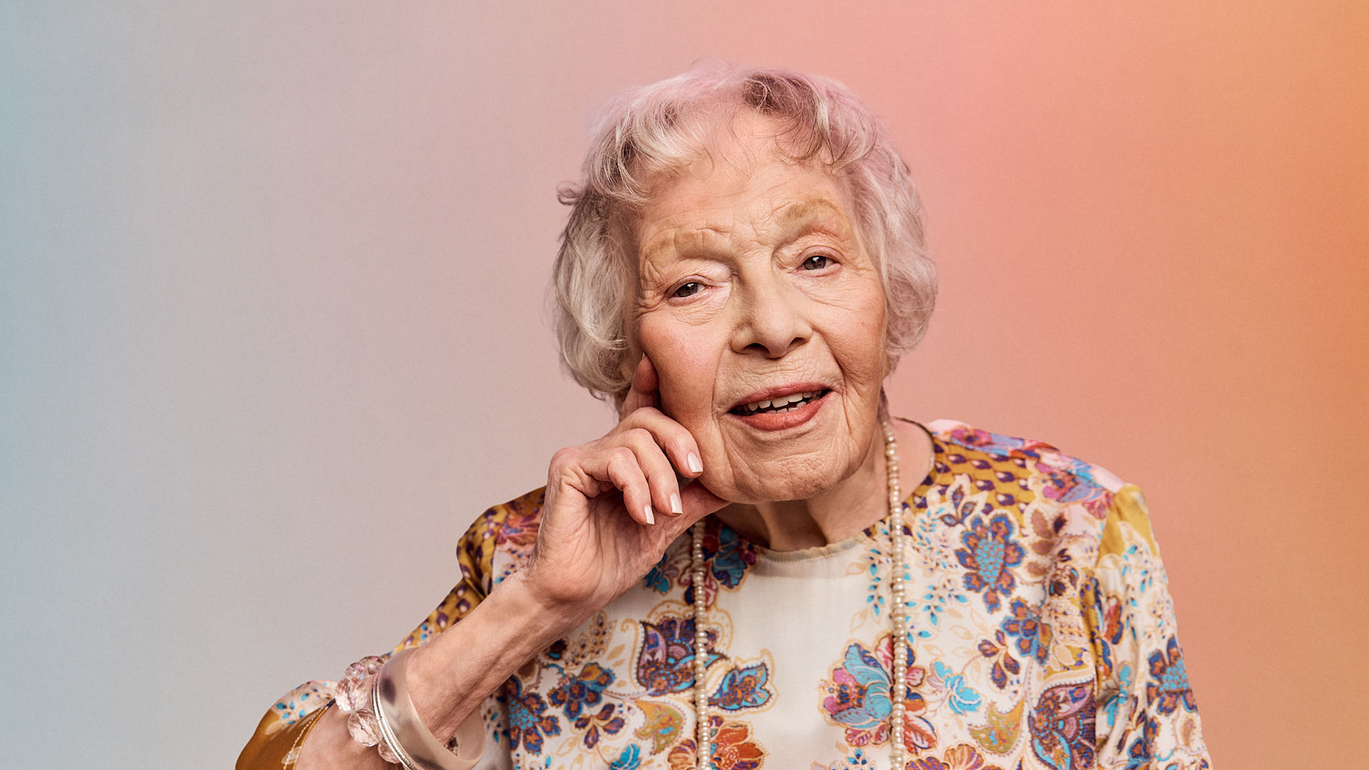 Older woman with short white hair, wearing a floral blouse, smiling gently, with her hand resting on her cheek against a soft gradient background.