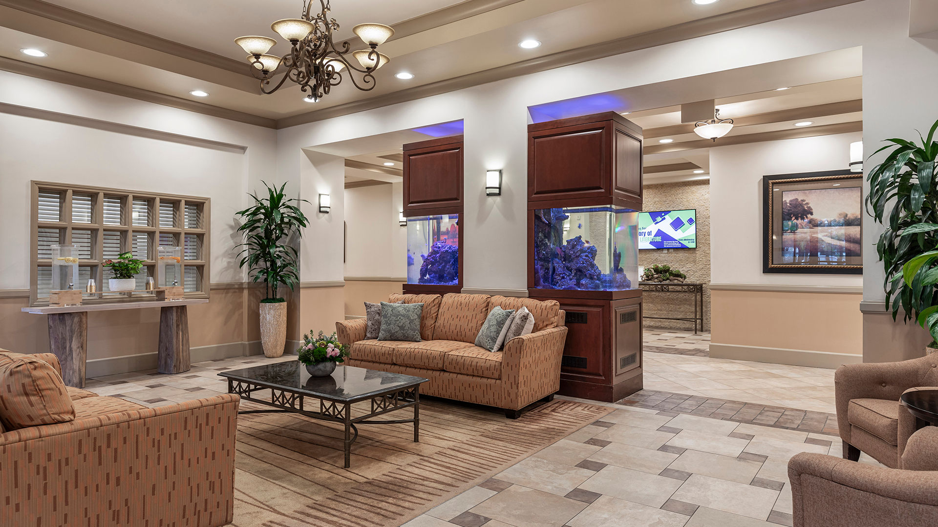 Elegant hotel lobby with sofas, a coffee table, large plants, and a decorative fish tank. Neutral tones and soft lighting create a welcoming ambiance.