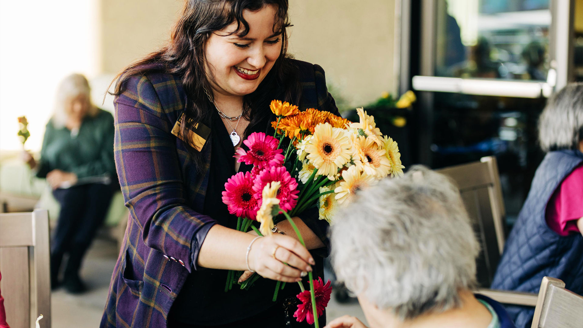 A woman smiling as she hands colorful flowers to an older person in an outdoor setting.