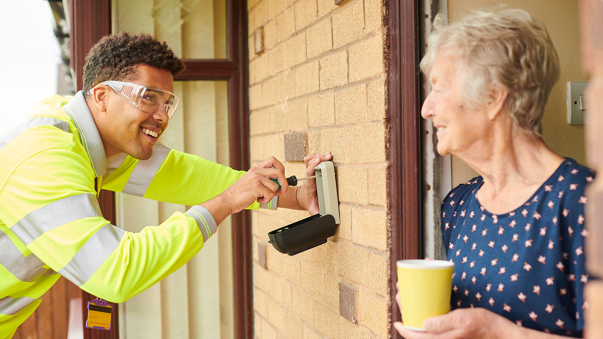 Maintenance man installs home alert system while an older woman holding coffee smiles at him from her doorway