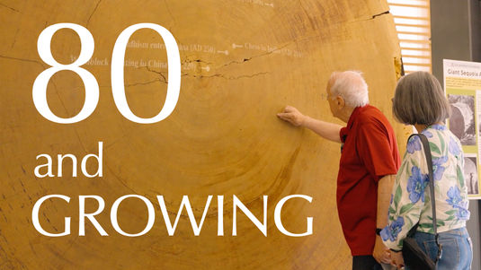 "80 and Growing" video thumbnail text overly of an image of two seniors exploring a museum exhibit