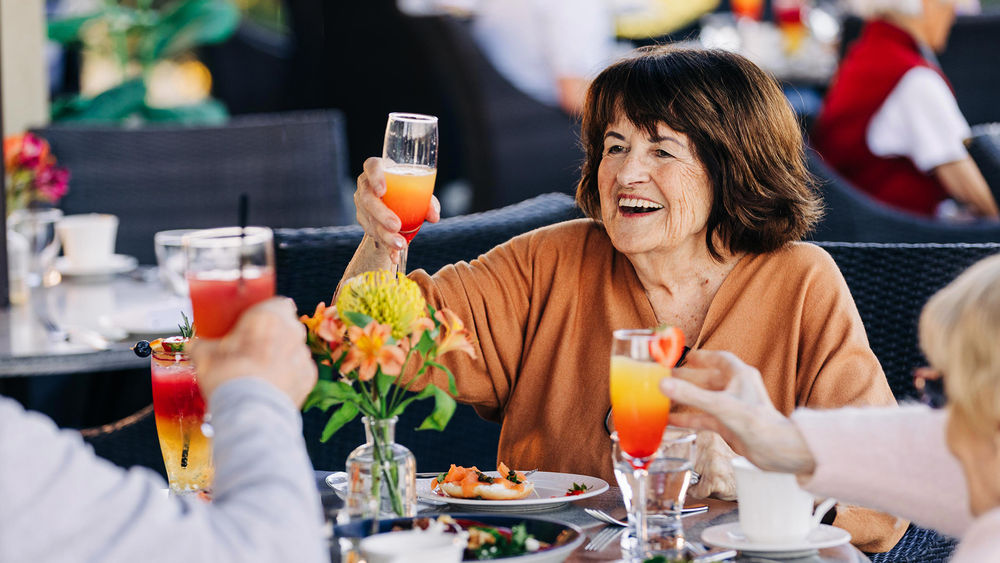 Older woman smiling and raising a glass at an outdoor restaurant table with friends, surrounded by flowers and plates of food.