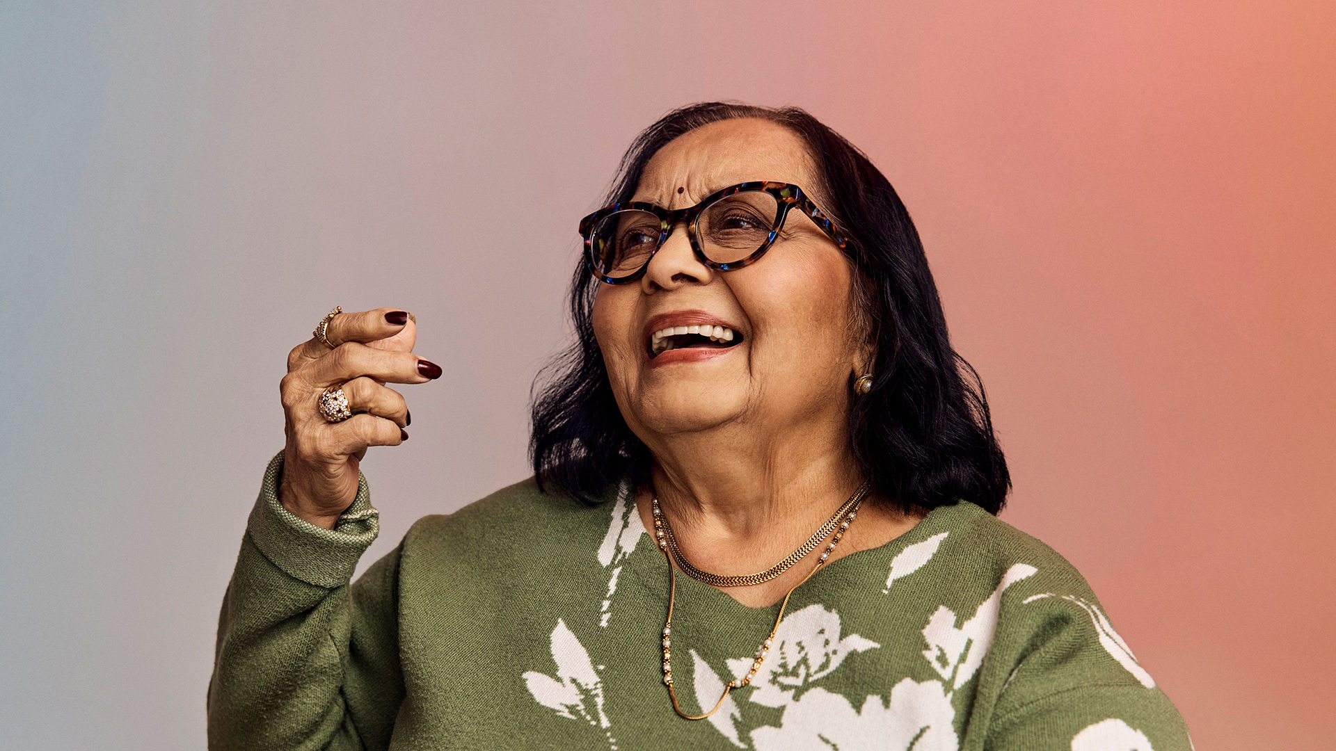 Smiling older woman with glasses, wearing a green floral sweater, holding a hand up against a gradient background.