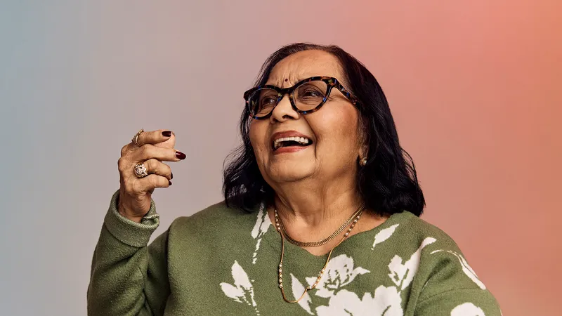 Smiling older woman with glasses, wearing a green floral sweater, holding a hand up against a gradient background.