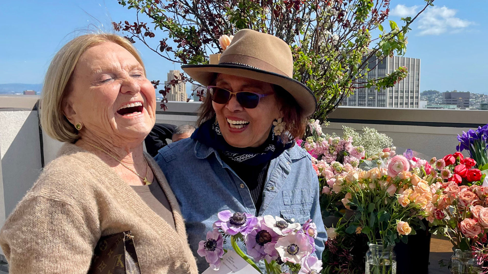 Two women laughing joyfully on a rooftop, surrounded by colorful flowers. One holds a bouquet, and the other wears a hat and sunglasses.
