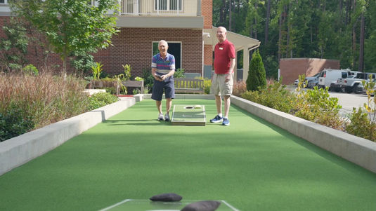 Two men playing cornhole outdoors on a sunny day, with a brick building and greenery in the background.