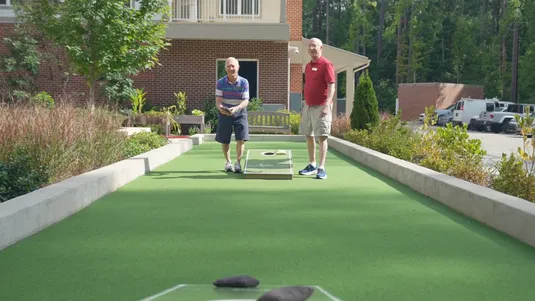 Two men playing cornhole outdoors on a sunny day, with a brick building and greenery in the background.