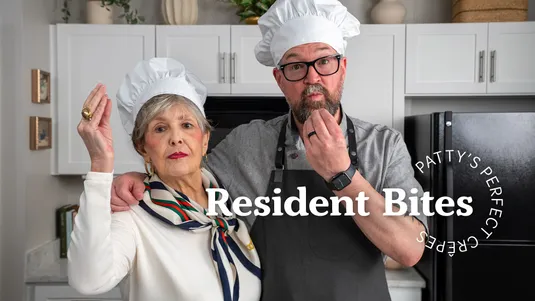 Two people wearing chef hats in a kitchen, posing playfully with hand gestures. Text overlay reads "Resident Bites Patty's Perfect Crepes."