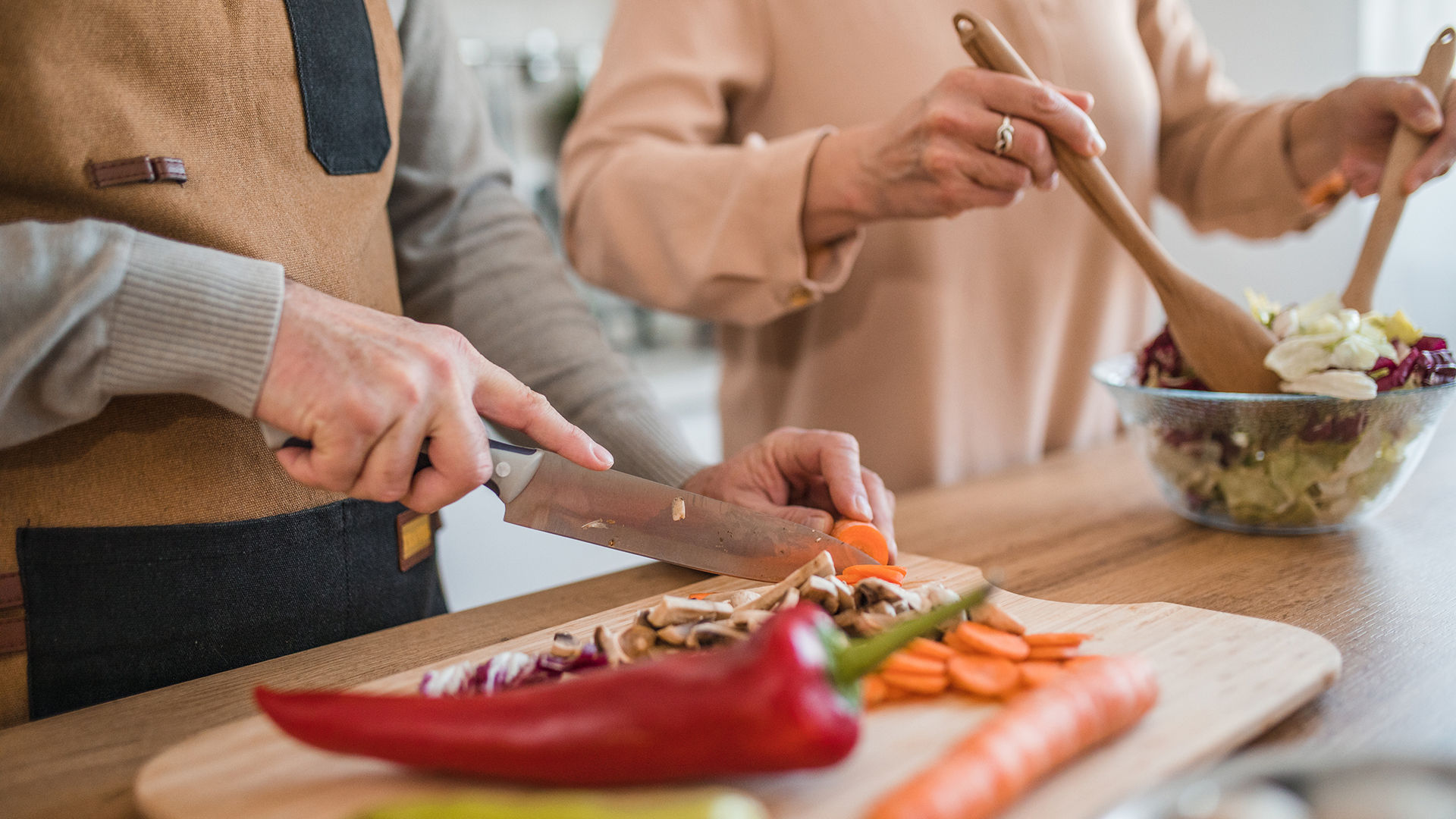 Two people preparing a meal; one slices vegetables on a cutting board, while the other tosses a salad in a bowl nearby.