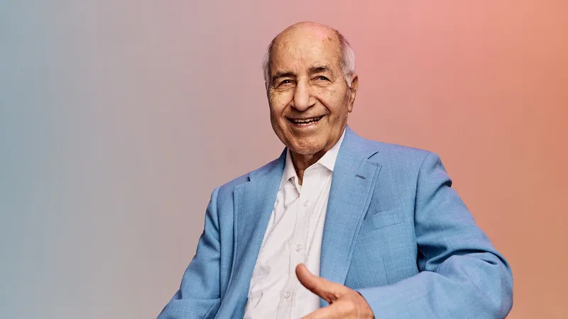 Older man smiling warmly, wearing a light blue suit and white shirt, with a soft gradient background.