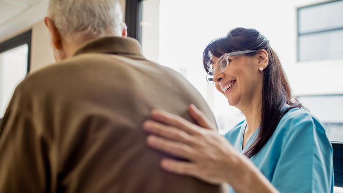 A caregiver in blue scrubs smiles and gently places a hand on an older man's back, offering comfort near a window.