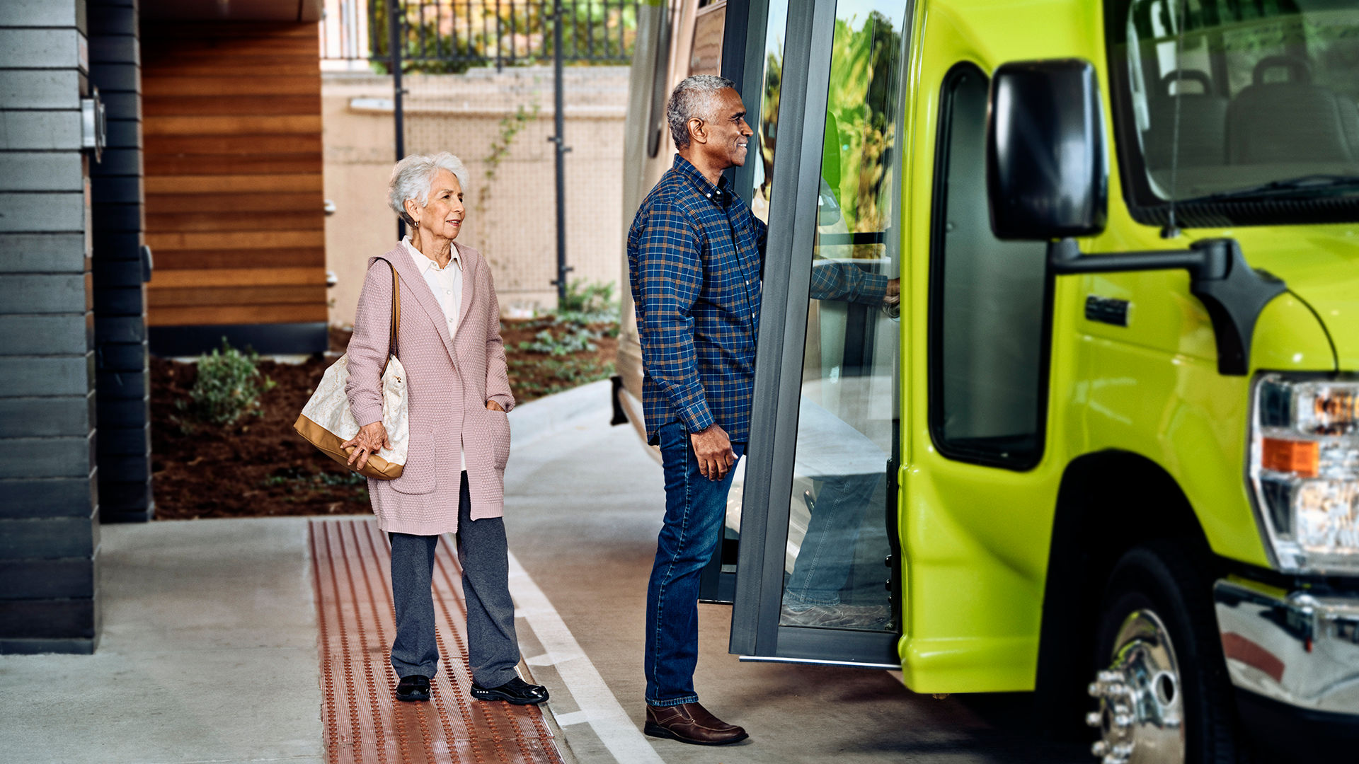 An older couple boards a bright green bus at a sheltered stop, with the woman holding a handbag and the man smiling.