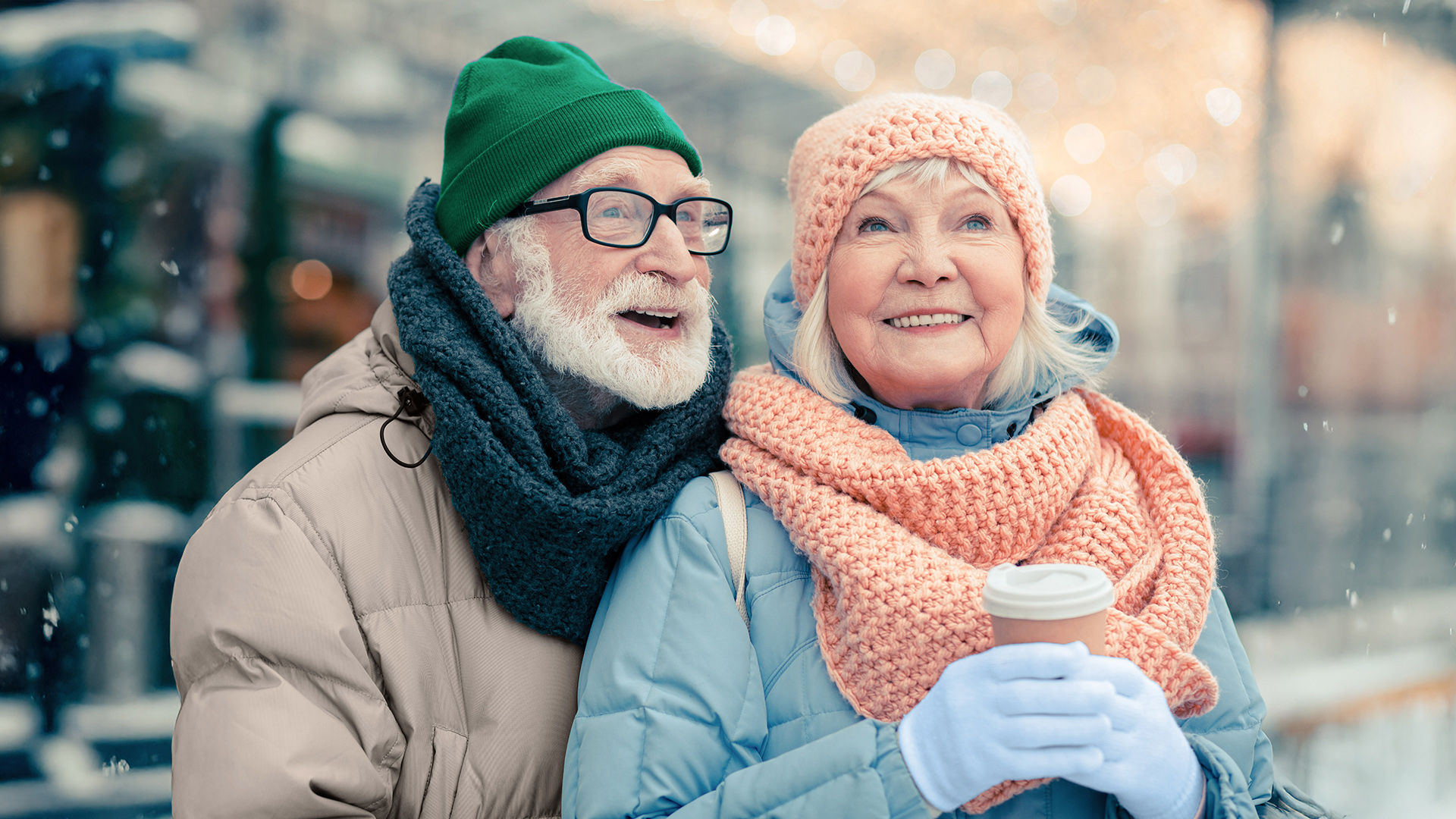 Smiling older couple in winter coats and knit hats; woman holds a takeaway coffee cup, both looking up amid falling snow and blurred lights.