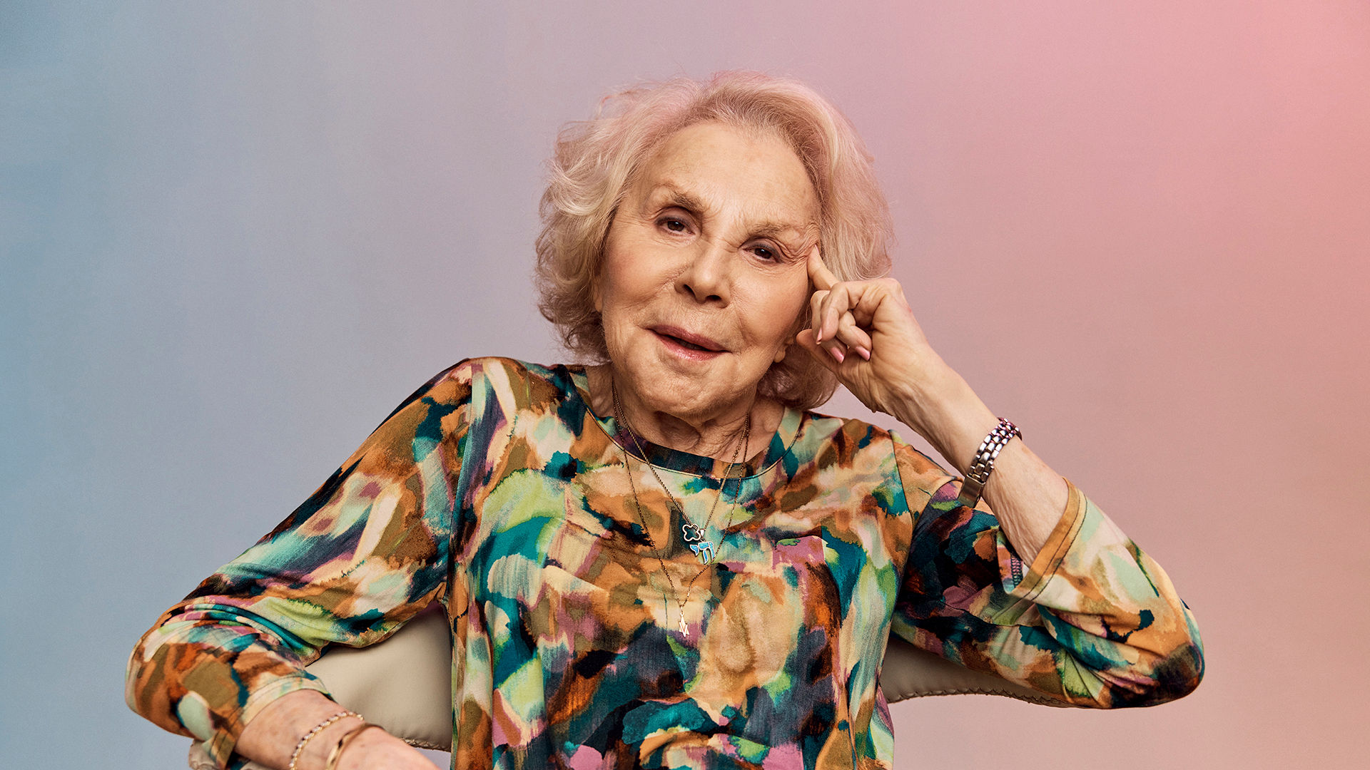 Older woman with blond hair, wearing a colorful blouse, sitting and smiling with her hand on her face against a gradient background.