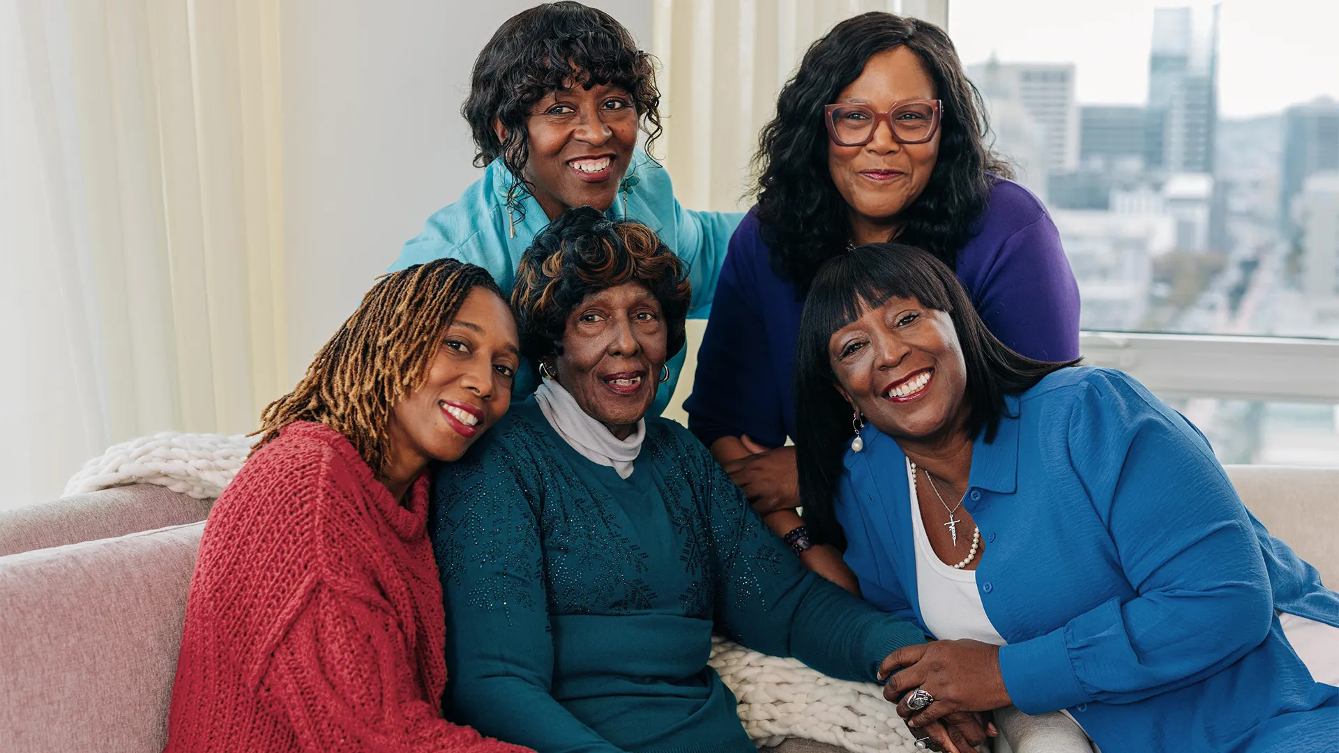 Five women of varying ages smile together in a cozy, bright room with a city view.