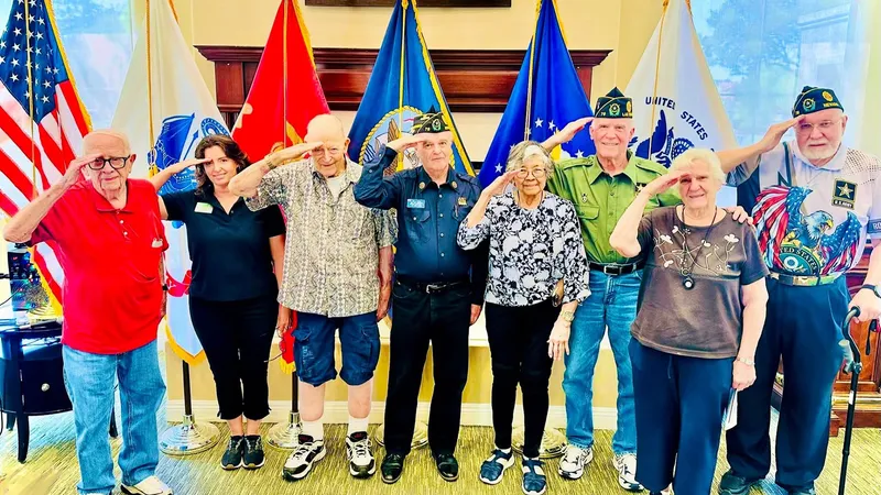 A group of elderly veterans and a woman stand saluting in front of various military flags indoors.