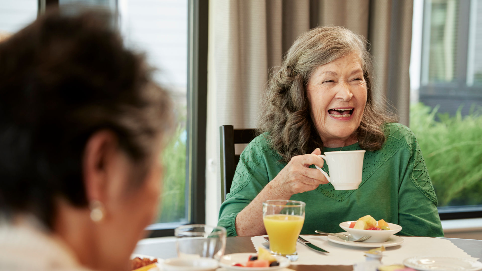 Senior woman in a green top laughing while holding a cup at a dining table with food and drinks, sitting near a window.