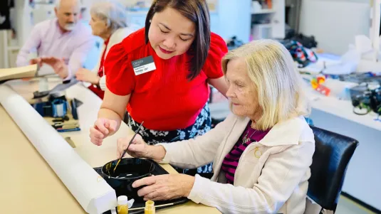 Senior resident in painting class being assisted by community staff.