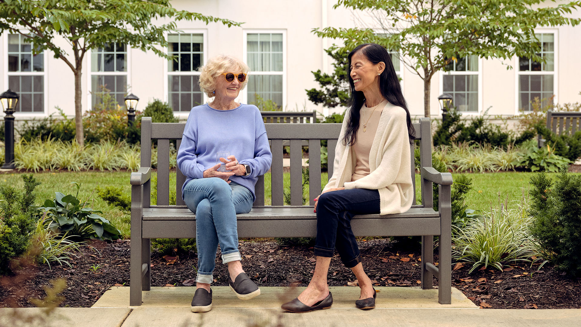 Two women sitting on a bench in a garden, smiling and chatting. One wears sunglasses; both are dressed casually.