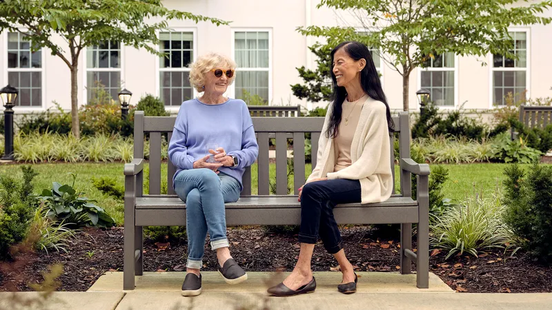 Two women sitting on a bench in a garden, smiling and chatting. One wears sunglasses; both are dressed casually.