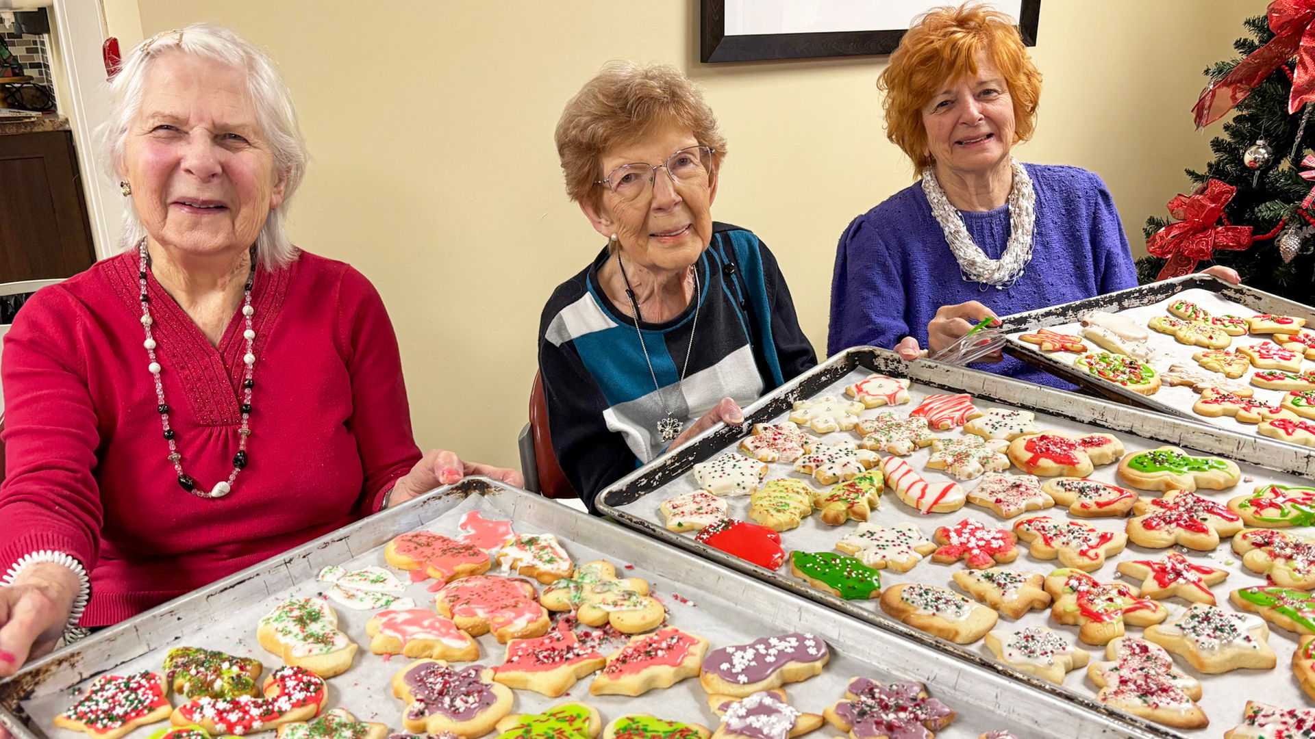 Three older women smile as they hold large baking trays filled with colorful, decorated holiday cookies. They sit indoors near a Christmas tree, proudly displaying cookies shaped like stars, trees, and other festive designs with red, green, white, and pink icing.