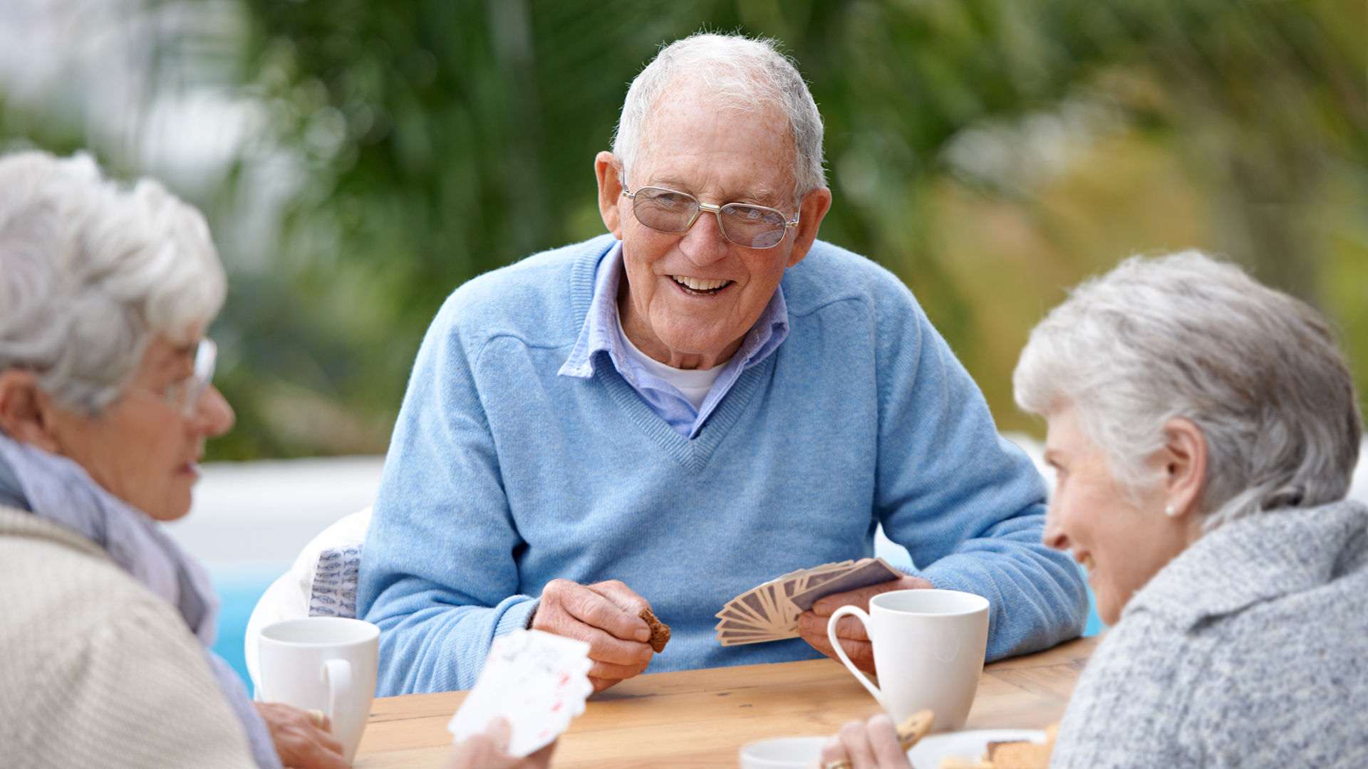 Older adult friends sitting outdoors, smiling and playing cards at a wooden table with coffee mugs, surrounded by greenery.