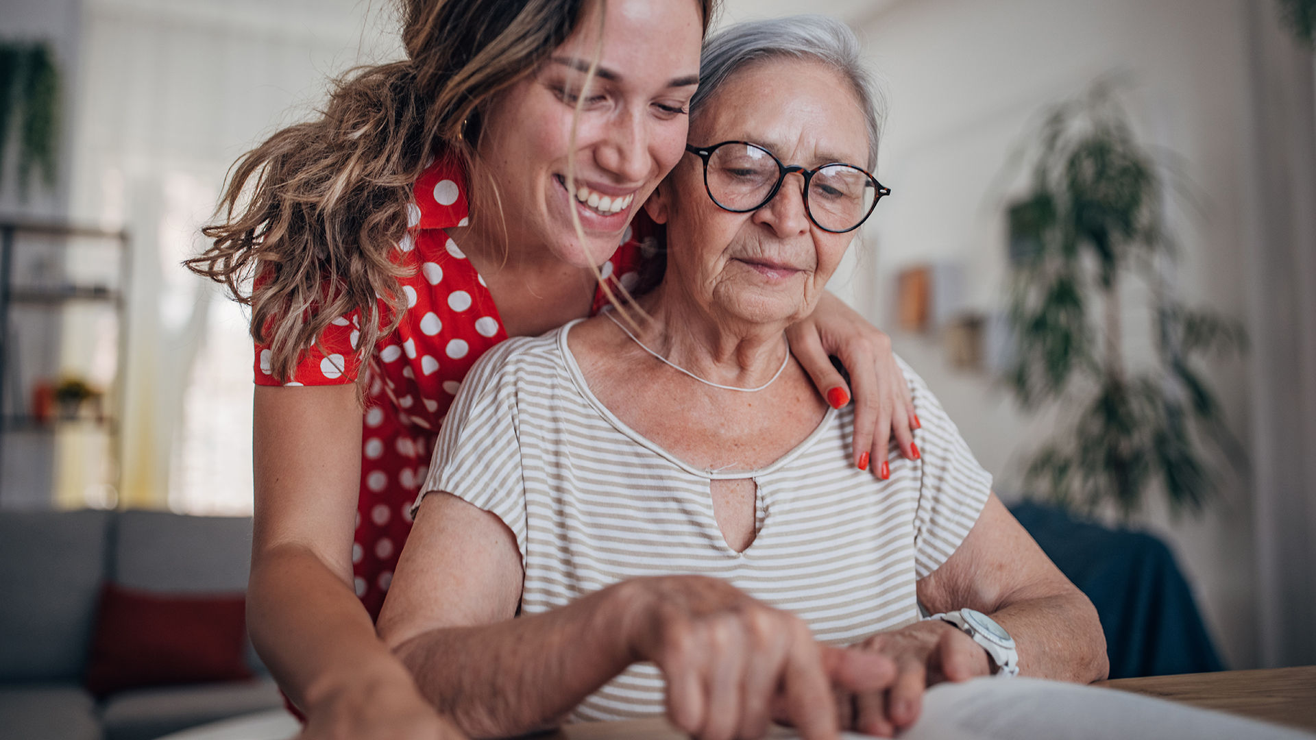Senior resident sitting at table pointing to writing in a book with adult daughter over her shoulder smiling.