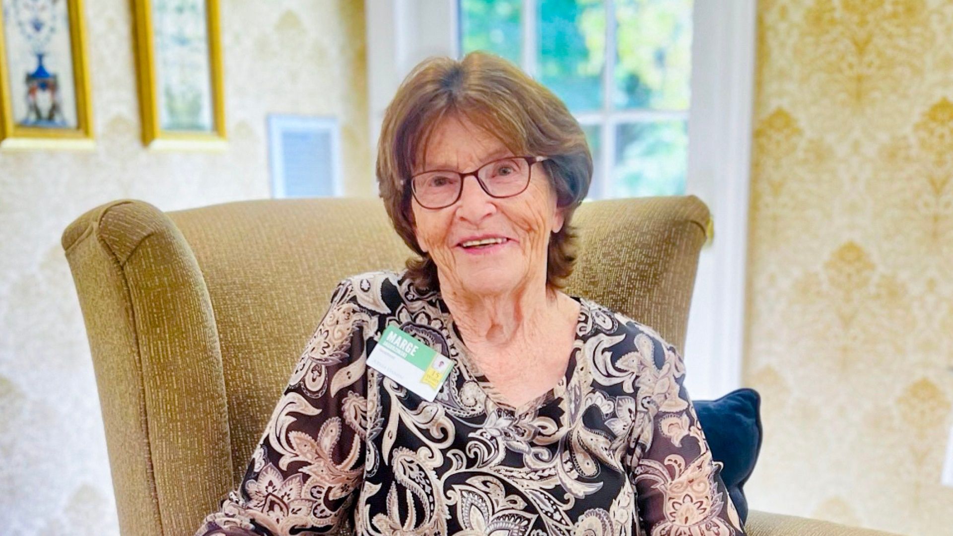 Marge, Atria Darien employee, smiles for the camera while seated in a tan chair.