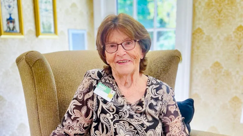 Marge, Atria Darien employee, smiles for the camera while seated in a tan chair.
