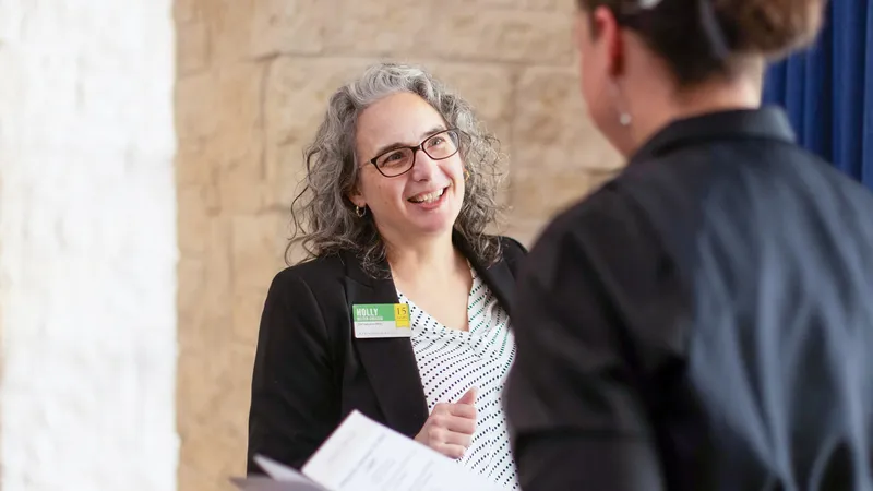 Woman with curly gray hair and glasses smiling, engaged in conversation with another person, holding a document in a bright room.