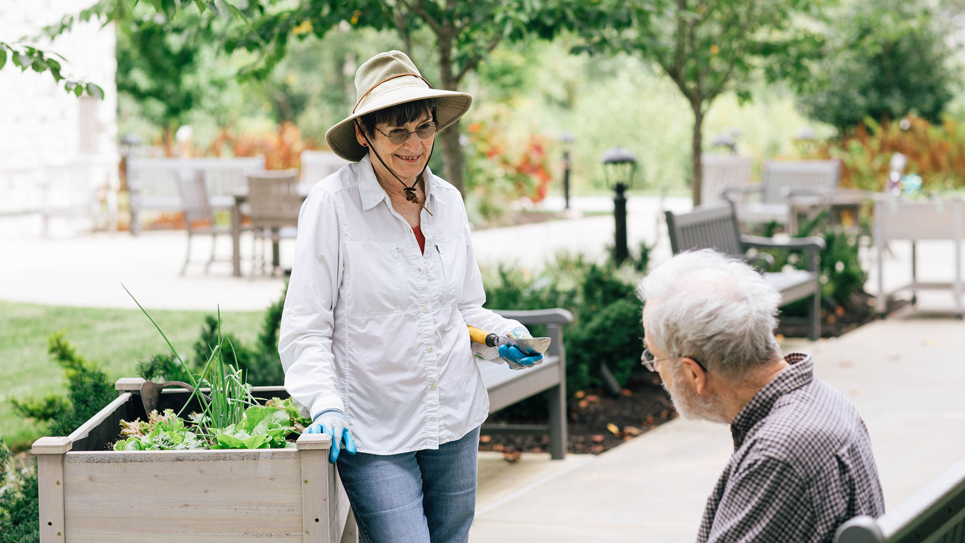 Older woman pauses gardening to talk to older man on bench