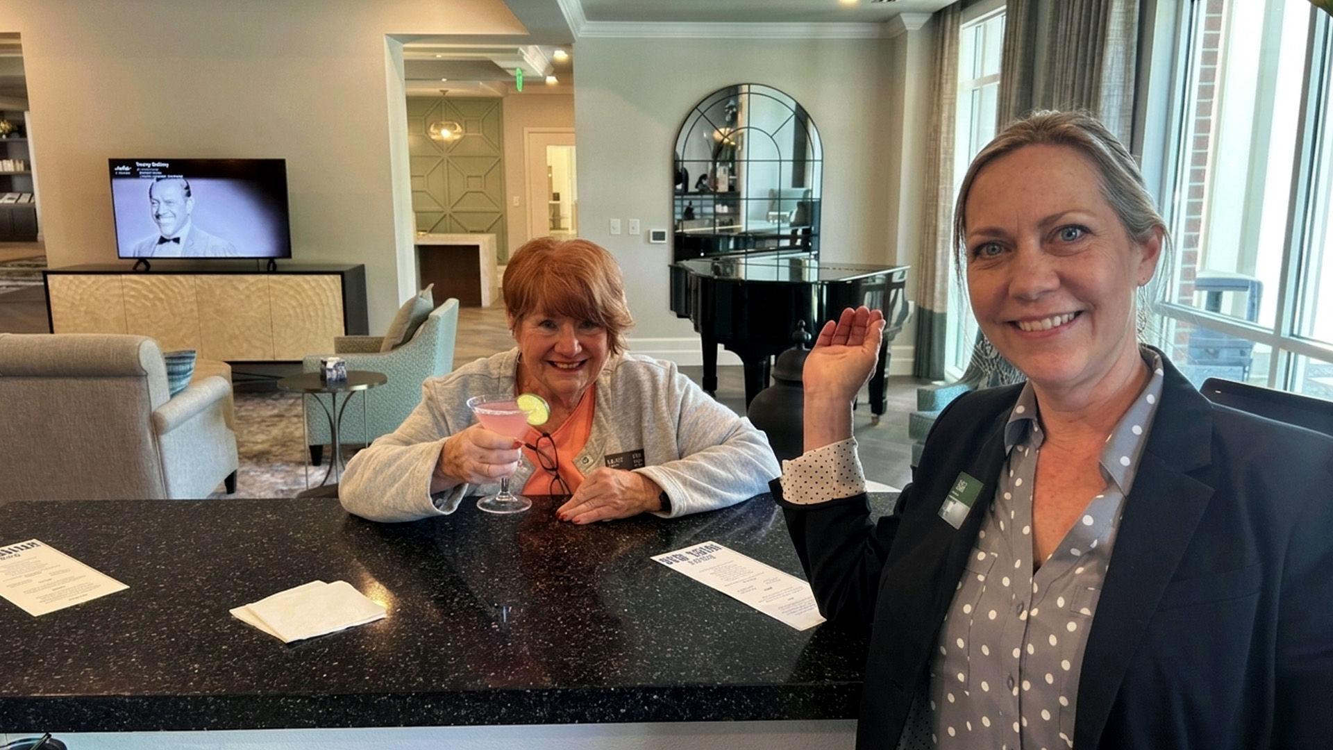 Two women at a bar counter; one holds a pink cocktail and smiles, while the other gestures with a smile. A grand piano is in the background.