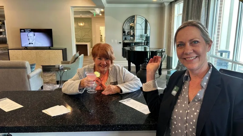 Two women at a bar counter; one holds a pink cocktail and smiles, while the other gestures with a smile. A grand piano is in the background.