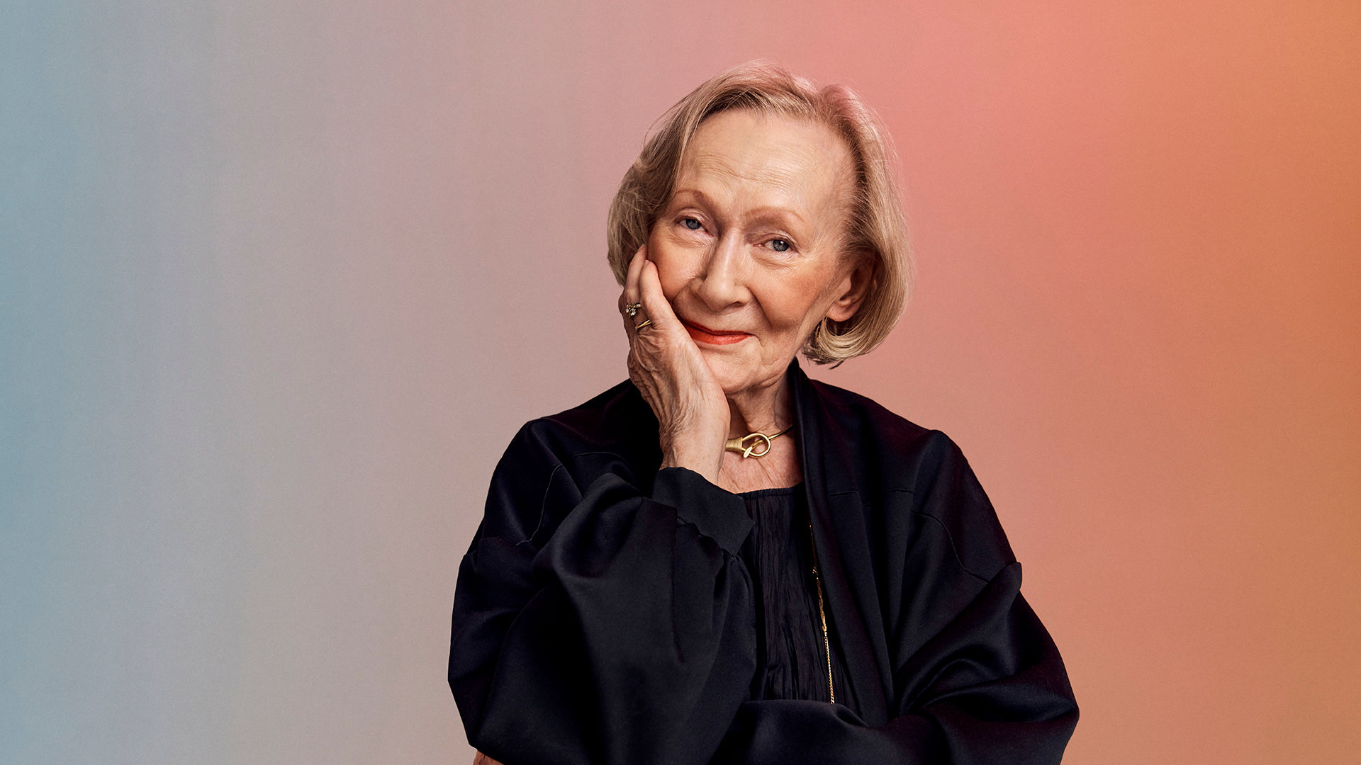 Older adult woman with short blonde hair smiles gently, resting her chin on her hand against a soft gradient background.