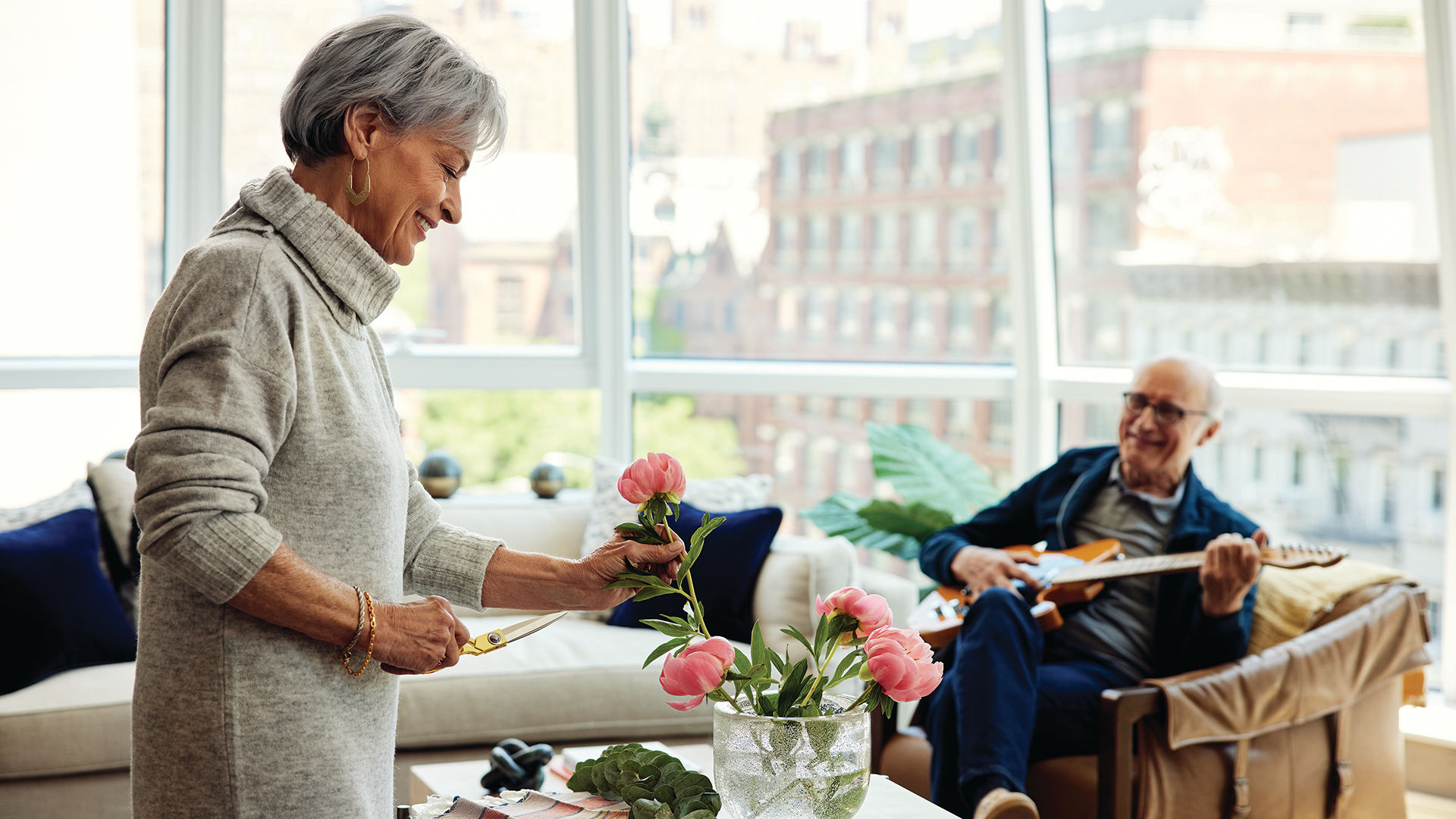 Coterie resident, an elderly woman, arranges flowers while smiling man plays guitar in a bright, modern living room with large windows.