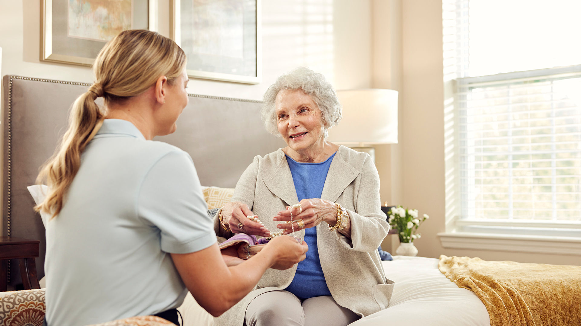 A caregiver helps an older woman in a bedroom. Both are smiling; the room is bright with a bed, pillows, and a window in the background.