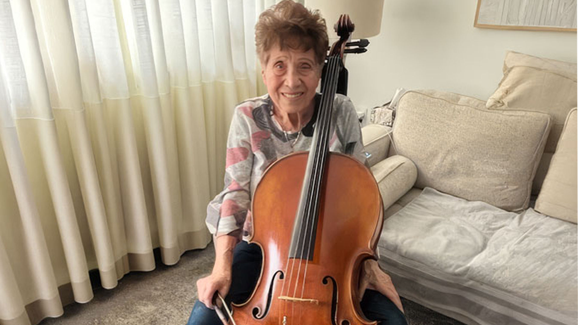 A senior woman smiling while sitting on a couch, holding a cello with a bow in her hand, in a room with white curtains.