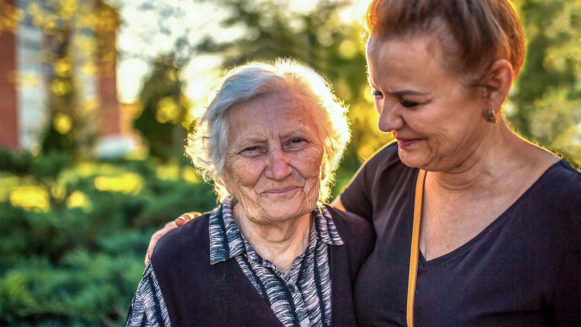 Adult daughter with arm around senior mother outdoors on a walk.