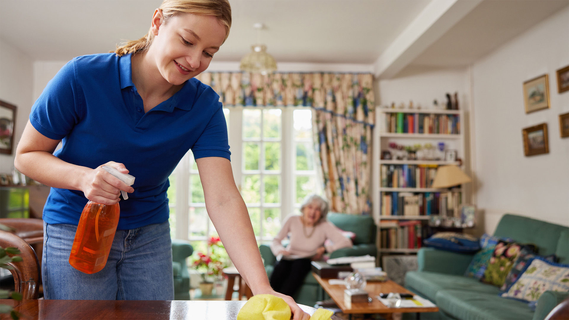 Housekeeper cleaning a senior resident's apartment with resident reading in a chair in the background.