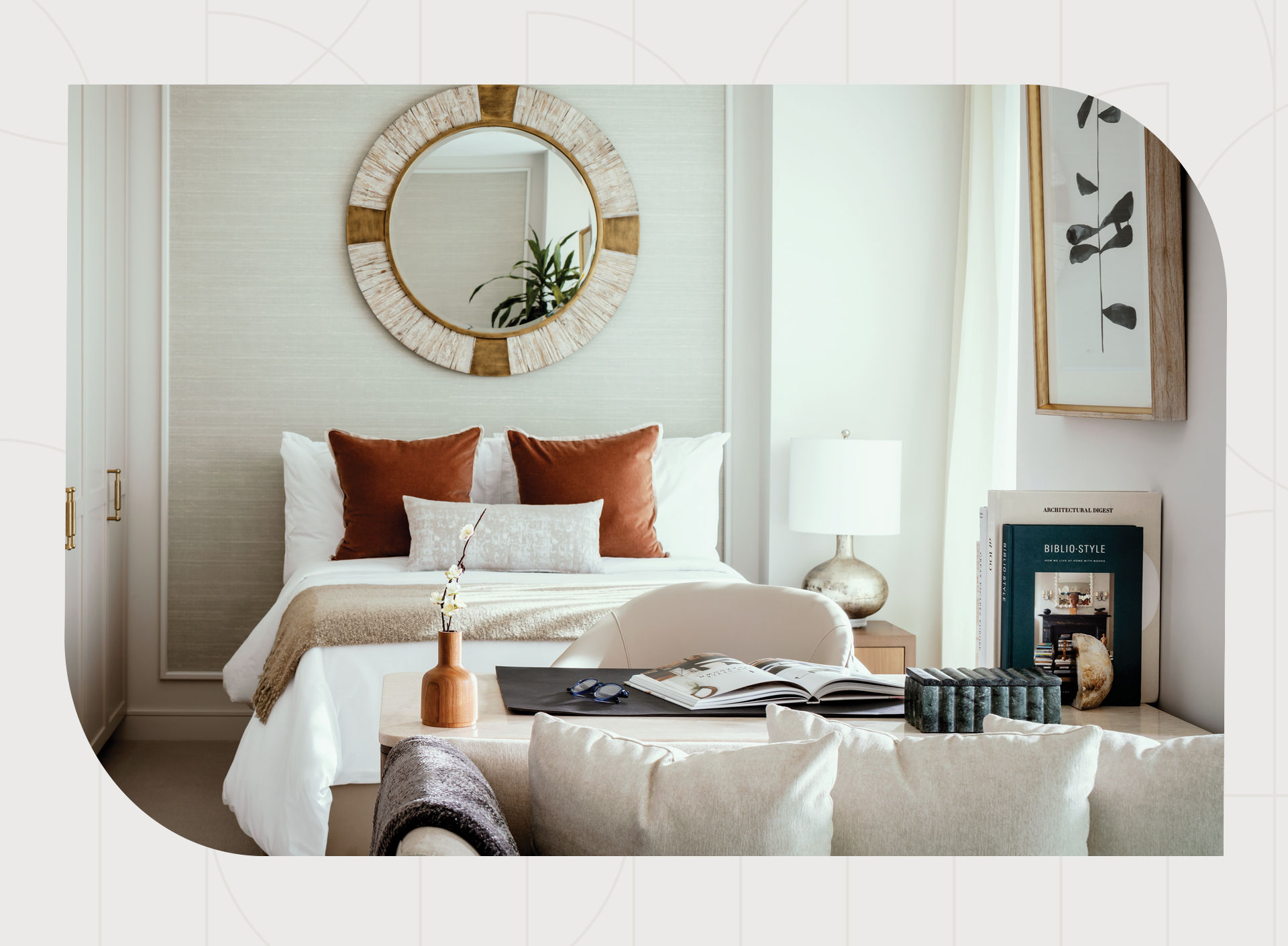 A cozy bedroom with a bed, rust-colored pillows, round mirror, lamps, books, and decorative items on a wooden table with a soft armchair.