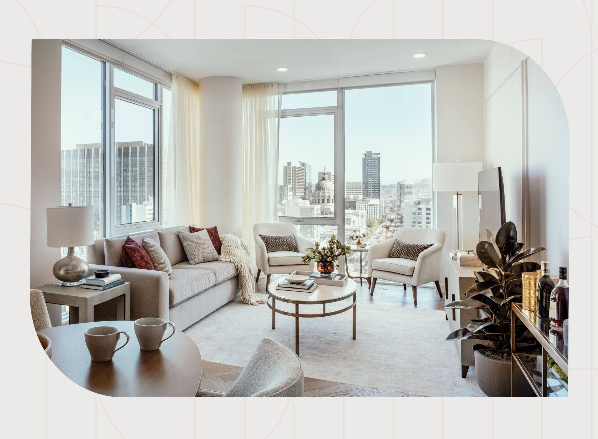 Bright modern living room with large windows, city view, neutral furniture, and decor, including a sofa, armchairs, and a potted plant.