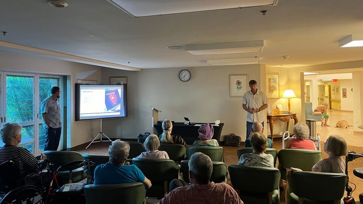 Residents seated in a warmly lit room watching two presenters speak near a projection screen displaying a scientific diagram