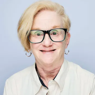 Smiling person with short blonde hair and glasses, wearing a light-colored shirt against a plain background.