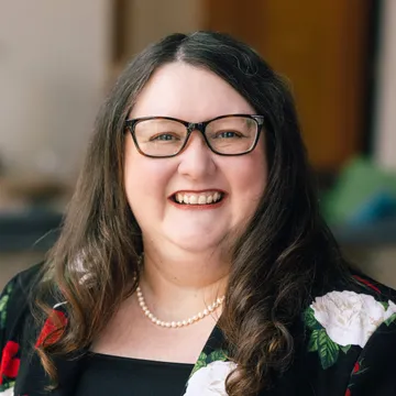 Smiling woman with long brown hair, wearing glasses, a floral-patterned top, and a pearl necklace against a blurred background.