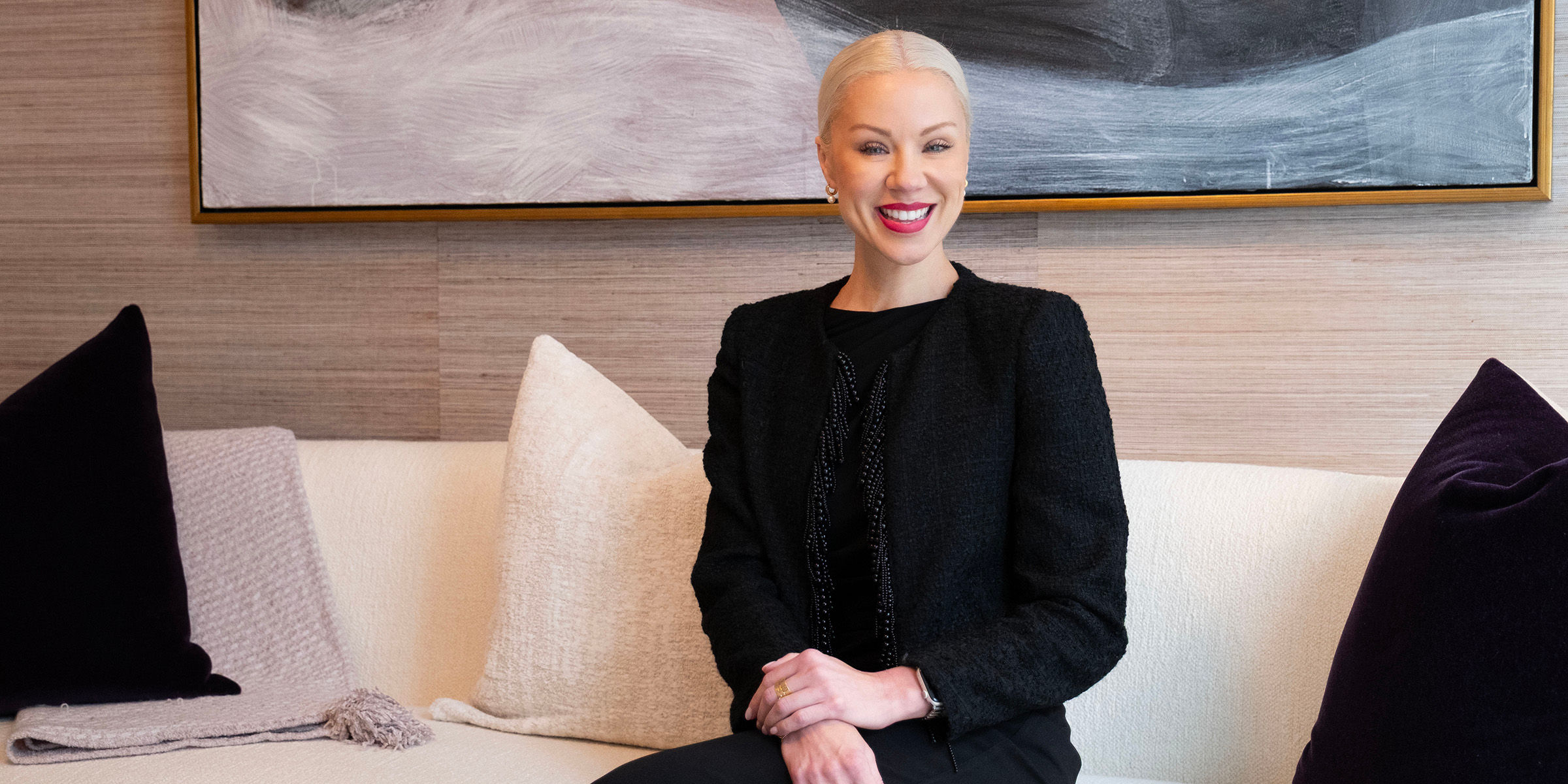 A woman with blonde hair in a black outfit sits on a white couch, smiling. A large abstract painting hangs on the wall behind her.