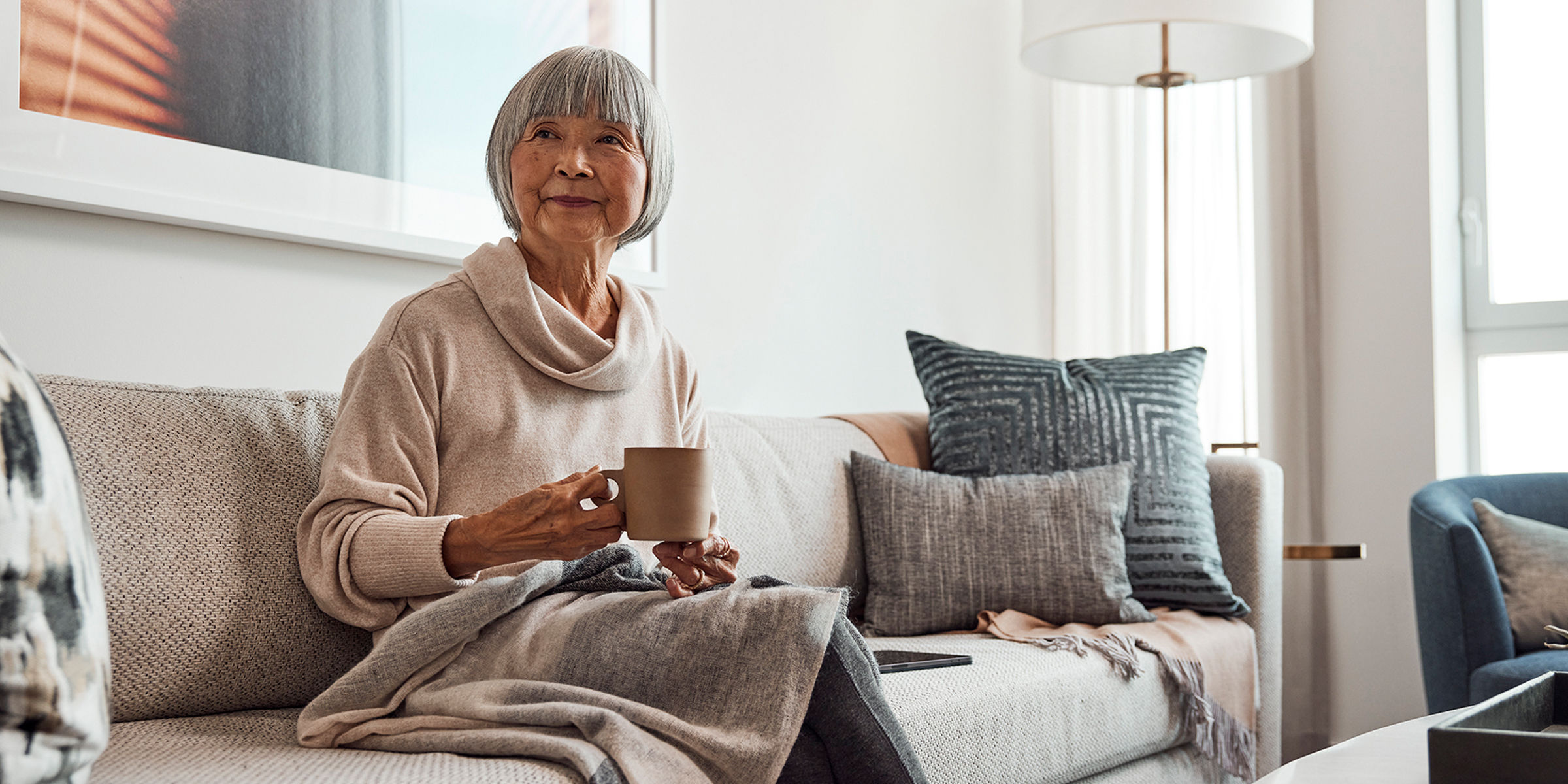 Stylish older woman sits on her couch; she is covered with a soft blanket in her lap and enjoys a warm cup of tea.