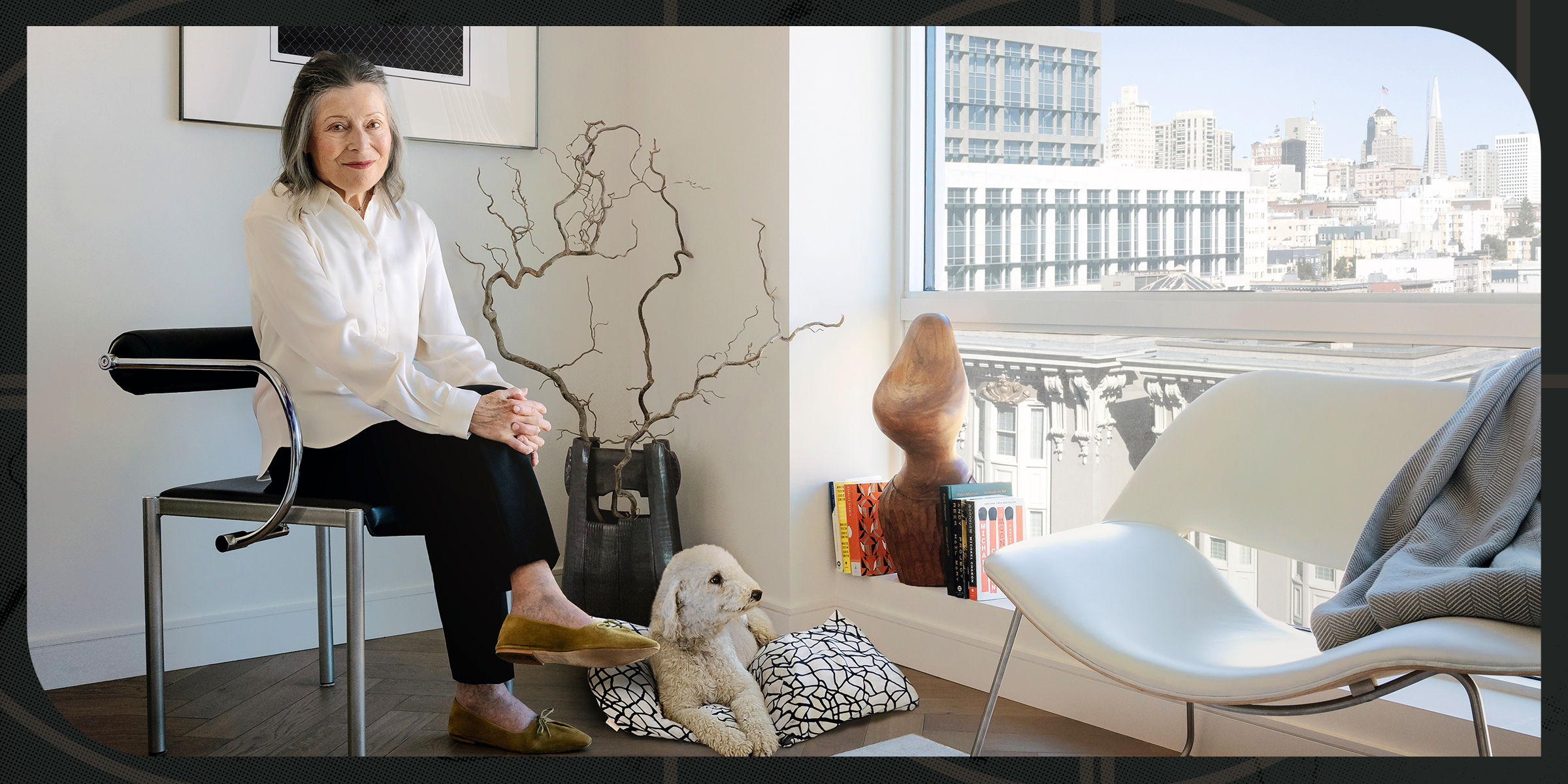 A woman in a white blouse sits on a chair in a modern room with large windows, city view, abstract decor, and a poodle-like dog on the floor.